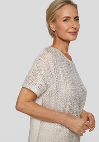 Silbernes gestricktes Top mit kurzen Ärmeln und einem glatten beigen Rücken. Das Material hat eine schimmernde Textur, die einen schichtartigen Effekt erzeugt.