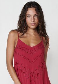 Rotes ärmelloses Kleid mit V-Ausschnitt, verziert mit Perlenstickereien in Chevron-Muster und Zickzackdetails am Saum. Weicher Stoff. 