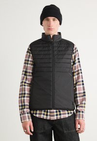 Colete puffer preto com gola alta, fecho de zíper e textura acolchoada, sobre uma camisa xadrez multicolorida com mangas em contrate.
