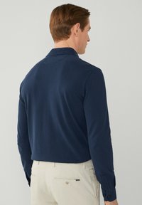 Camicia a maniche lunghe blu navy con colletto piatto, tessuto liscio e polsini abbottonati, abbinata a pantaloni beige chiaro con tasca posteriore.