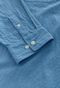 Poignet de chemise bleu à manches longues avec deux boutons blancs sur un tissu texturé, mettant en valeur les détails de couture et le placement des boutons.