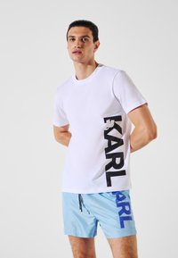 T-shirt en coton blanc avec le logo "KARL" noir sur le côté. Porté avec un short de bain bleu clair présentant un imprimé vertical "KARL" en bleu.
