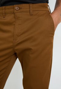 Brown cotton trousers featuring a smooth texture, a button closure, and side pockets. The stitching is consistent, emphasizing the fitted design.
