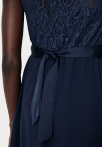 Tums zilas kleita ar mežģīņu augšu, kas dekorēta ar ziedu rakstiem, gluda satīna josta un pievilkšanas bante aizmugurē.