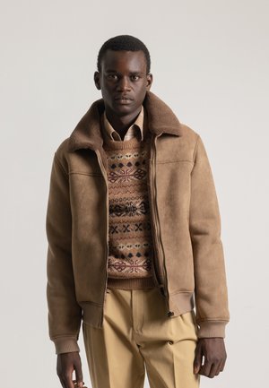 Chaqueta de piel de oveja marrón con cuello de felpa, cierre de cremallera, combinada con un suéter estampado y pantalones beige. Los materiales texturizados son visibles.