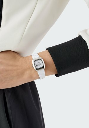 Ceas cu curea de cauciuc albă, cu o carcasă pătrată argintie și un cadran minimalist, cu markeri de oră subtili. Este purtat la încheietura mâinii, pe un mânecă neagră.