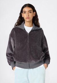 Veste à capuche en velours zippée de couleur gris foncé avec un col montant, des poignets côtelés et des poches latérales. Texture douce, coupe décontractée, associée à une chemise blanche en dessous.