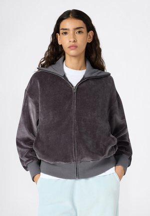 Fluwelen zip-up hoodie in donkergrijs met een hoge kraag, geribde boorden en zijzakken. Zachte textuur, losse fit, gecombineerd met een wit shirt eronder.