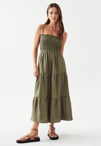 Olivegrünes Maxikleid aus weichem Stoff mit gerafftem Oberteil, Rüschenausschnitt und gestuftem Rock; kombiniert mit braunen Riemchensandalen.