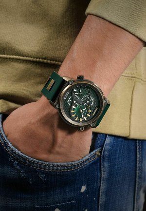 Grüne Uhr mit strukturiertem Ziffernblatt und Gehäuse in Messingoptik, kombiniert mit einem grünen Silikonarmband. Sichtbar an einem Handgelenk in der Nähe von Denimstoff.