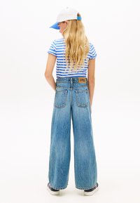 Φαρδιά μπλε τζιν παντελόνια με ανοιχτό πλύσιμο, με καφέ δερμάτινο λογότυπο. Συνδυάζονται με μπλε και λευκό ριγέ μπλουζάκι και καπέλο.