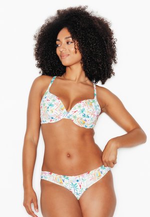 Donna con capelli ricci naturali che indossa un bikini colorato con stampa tropicale, sorridente e che guarda verso sinistra su sfondo bianco.