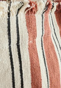 Tissu tissé présentant des rayures alternées en terre cuite, crème et noir. Les bords ont des franges effilochées. Tissu doux et texturé.