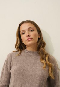 Beżowy sweter prążkowany z wysokim, okrągłym dekoltem, wykonany z miękkiego materiału. Modelka ma długie, falujące włosy i stoi na neutralnym tle.