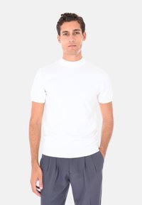 Giovane uomo con capelli corti e ricci, che indossa una semplice camicia bianca a maniche corte e pantaloni grigi scuri con pieghe, mani in tasca, sfondo neutro.