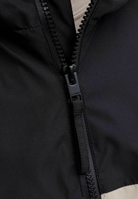 Veste noire avec un col montant, dotée d'une fermeture éclair centrale et d'un panneau beige contrastant en bas. Texture lisse, matériau léger.