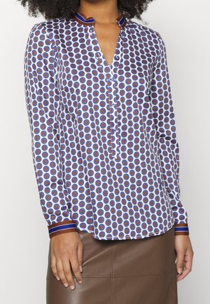 Donna che indossa una camicetta a maniche lunghe con motivo a cerchi blu e marrone, polsini e collo rifiniti in blu, abbinata a una gonna di pelle marrone.