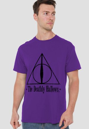 Man wearing a purple T-shirt with a black Deathly Hallows symbol and text, looking slightly to his right against a plain background.