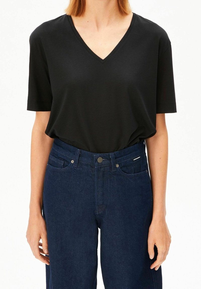 Personne portant un t-shirt noir à col en V avec manches courtes, rentré dans un jean bleu foncé taille haute, debout les bras relâchés.