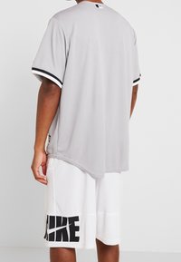Jersey atlético gris de manga corta, con acentos negros en los puños y dobladillo. Lleva pantalones cortos blancos que tienen un logo negro.