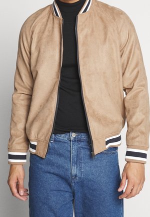 Personne portant une veste bomber en daim beige avec des poignets et un col rayés blancs et noirs, un col roulé noir et un jean bleu.