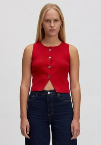 Frau mit langen blonden Haaren, die ein ärmelloses rotes Knopfhemd und dunkelblaue Jeans trägt, steht vor einem schlichten hellen Hintergrund.