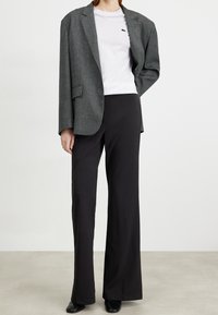 Šedý herringbone blazer se strukturovaným střihem, doplněný bílým tričkem s krátkým rukávem. Černé kalhoty s širokými nohavicemi a černé boty dokončují vzhled.