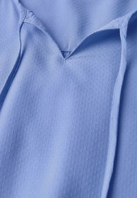 Tessuto blu chiaro con un motivo a pois testurizzato. Presenta un collo a V con chiusura a lacci e una morbida caduta. Adatto per un abbigliamento casual.