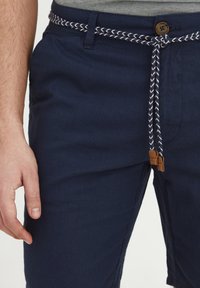 Navy blue shorts with a textured fabric, featuring a woven belt in white and navy, and brown leather accents at the ends of the belt ties.
