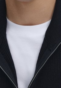 Svart stickad zip-up-jacka med ribbad textur, lager på lager över en vit T-shirt med rundad halsringning. Tydlig dragkedjedetalj längs halslinjen.