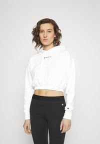 Felpa bianca corta con cappuccio regolabile e orlo elastico, presenta il logo "Champion" in nero. Abbinata a leggings neri.