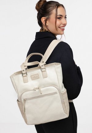 Jonge vrouw met bruin haar in een knot, gekleed in een zwarte jas en een beige rugzak met een voorvak met rits, die glimlacht terwijl ze opzij kijkt.