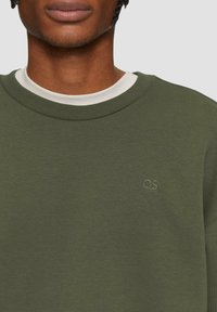 Olivgrüner Sweatshirt aus weichem Stoff mit rundem Halsausschnitt. Verfügt über ein dezentes, in derselben Farbe auf der Brust gesticktes "QS"-Logo.