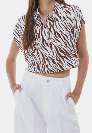 Kvinde iført en brun og hvid zebra-stribet ærmeløs cropped skjorte med knapper og hvide bukser med høj talje med hænderne i lommerne.