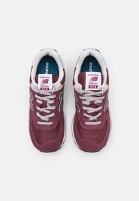 Burgundi színű textil sportcipők szürke fűzőkkel, hálós és velúr részletekkel, fehér középtalppal, valamint "New Balance Classic 574" felirattal a nyelvén.