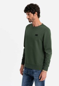 Felpa verde in cotone misto, con collo rotondo, maniche lunghe e un'etichetta logo rettangolare sul petto. Indossata con jeans blu.