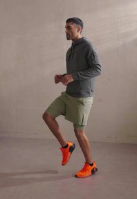 Grå hoodie, lysegrønne shorts og lysorange træningssko. Personen er i en gangbevægelse på en glat overflade.