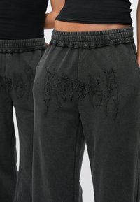 Mustat collegehousut, joissa on joustava vyötärönauha, sivutaskut sekä etupuolella teksturoitu, kirjailtu kirjainkuvio. Pehmeä, rento kangas.