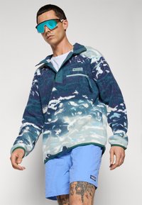 Mies, joka pitää päällään sinistä kuvioitua fleece-takkia, vaaleansinisiä shortseja, heijastavia sinisiä urheilulasia ja jota koristavat näkyvät jalkahautat, asetettuna yksinkertaista taustaa vasten.