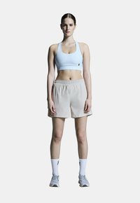 Helles blaues Sport-BH kombiniert mit beigen Shorts, die einen elastischen Bund haben. Das Model trägt weiße Socken und graue Sportschuhe.