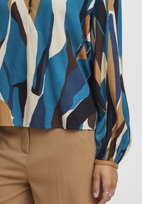Farbige Bluse mit abstrakten Mustern in Blau, Schwarz, Beige und Braun. Lange Ärmel mit elastischen Manschetten. Hergestellt aus glattem, leichtem Stoff.