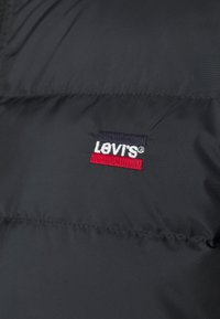 Svart vadderad jacka med en röd och marinblå broderad Levi's-logotyp på vänster bröst. Materialet har en slät, glänsande yta.