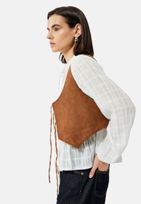 Gilet corto in suede marrone con orlo appuntito e lacci regolabili, indossato sopra una blusa bianca texturizzata. Il modello è rivolto a destra contro uno sfondo semplice.