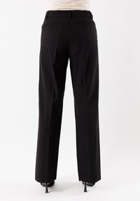 Pantaloni larghi neri con finitura liscia, passanti per la cintura, fronte plissettato e design sartoriale, mostrati di spalle con scarpe con tacco alto.