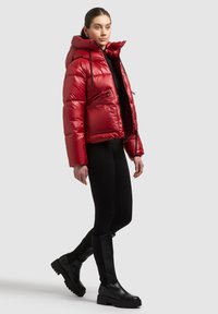 Sarkana pūku jaka ar kapuci, rāvējslēdzēju priekšpusē un sānu kabatām. Gluds, spīdīgs materiāls; īss piegriezums. Melnas legingi un ceļgalu augstie zābaki noslēdz kopējo tēlu.