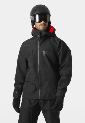 Mand iført sort skijakke, bukser, handsker, hjelm og goggles, poserende mod en ensartet lys baggrund.