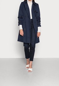 Marineblauer Trenchcoat mit doppelter Knopfleiste, Schulterklappen und tailliertem Gürtel. Getragen über einem weißen Rollkragenpullover und kombiniert mit dunkler Jeans und weißen Sandalen.