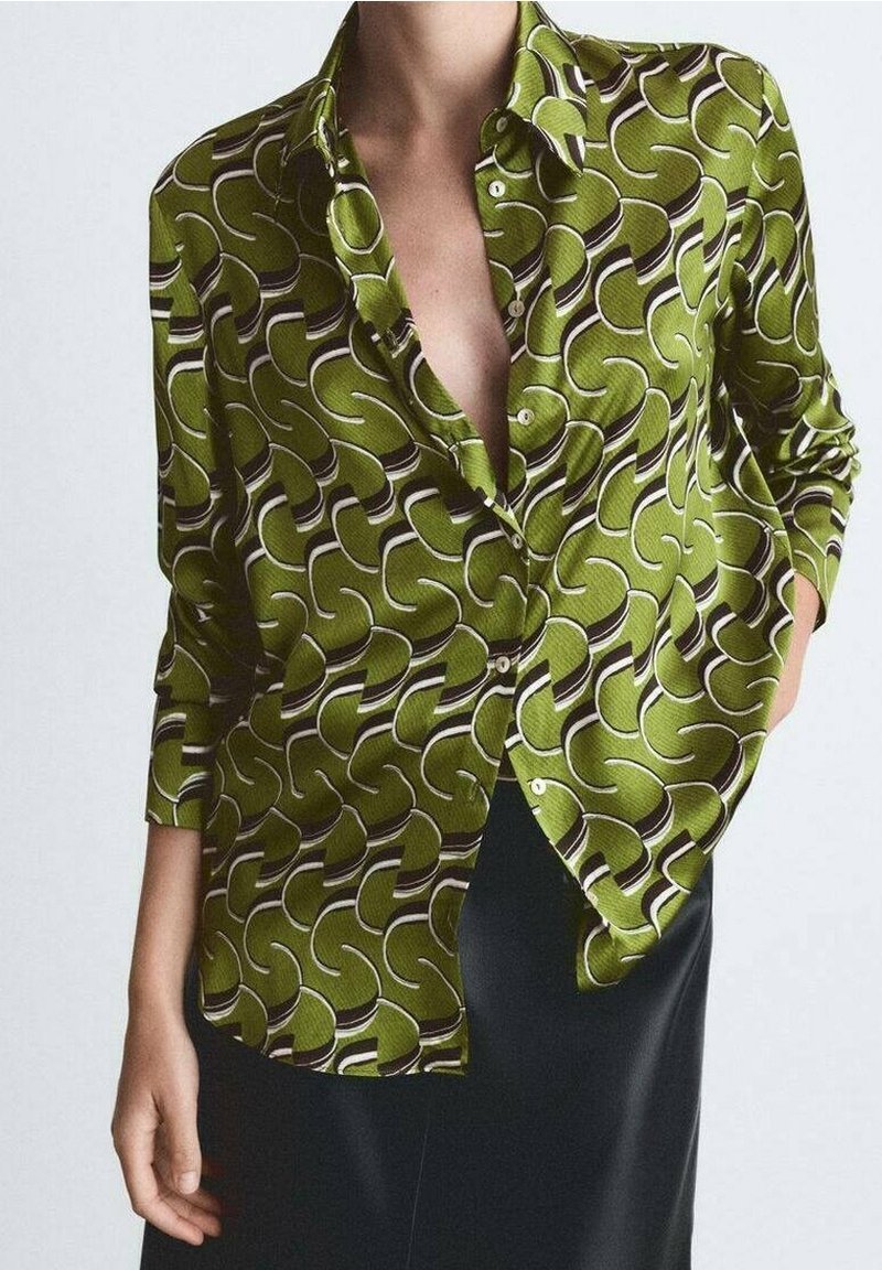 Personne portant une chemise verte à motifs déboutonnée à manches longues et une jupe noire, main dans la poche, sur un fond uni.
