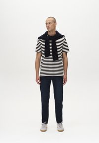 Camicia a righe a maniche corte in crema e blu navy, abbinata a jeans scuri e sneakers chiare. Maglione appoggiato sulle spalle, accessori minimi.