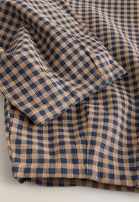 Gingham-mønstret stof i beige og marineblå, med en tekstureret vævning og synlige syninger langs kanterne samt et blødt fald.